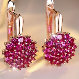 Puede incluir: Un par de pendientes de aro en oro rosa con un grupo circular de pequeñas piedras redondas de color rosa vibrante. Cada pendiente tiene un cierre de palanca articulado y un acabado metálico pulido que refleja la luz en las facetas.