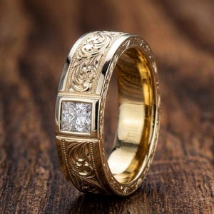 May include: A gold band ring featuring a square princess-cut clear gemstone set in the center. The outer surface of the gold band is decorated with intricate, engraved scrollwork patterns that extend around the circumference of the ring.