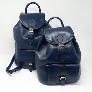 May include: Two navy blue leather backpacks with top handles, flap closures, and front pockets. The backpacks have adjustable straps and gold-colored buckles. One backpack is slightly larger than the other.