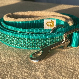 Puede incluir: Una correa para perro de color azul turquesa con un cierre plateado y una pequeña etiqueta blanca con letras doradas. La correa está decorada con diamantes de imitación verdes.