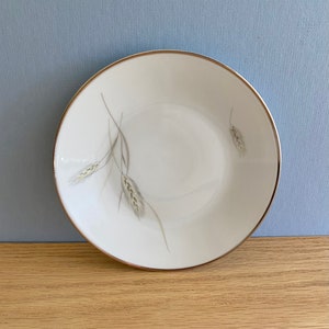 May include: A white ceramic bowl with a silver rim and a wheat design. The bowl is on a wooden surface.