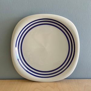 May include: A white ceramic plate with a slightly rounded shape and three blue stripes around the edge.