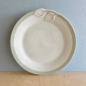 Może przedstawiać: Biały ceramiczny talerz z jasnozieloną obwódką i dekoracyjnym uchwytem. Talerz ma prosty, elegancki design ze srebrnym wykończeniem. Talerz leży na drewnianej powierzchni z niebieskim tłem.