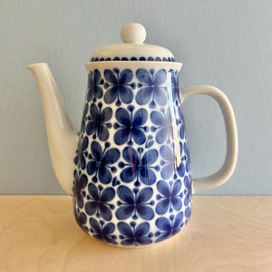Op de afbeelding: Een witte en blauwe keramische koffiepot met een deksel. De pot is versierd met een blauw bloemenpatroon. De handgreep en de tuit zijn wit. De pot staat op een licht houten oppervlak.