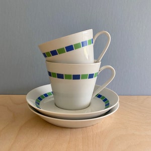 May include: Two white ceramic coffee cups with blue and green geometric patterns stacked on top of each other, with a matching saucer underneath.