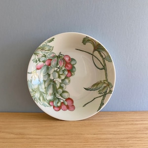 Peut inclure: Un bol rond en céramique blanc cassé avec un motif de raisin peint. Les raisins sont un mélange de vert et de rose, avec des feuilles et des vignes vertes. Le bol est posé sur une surface en bois sur fond bleu clair.