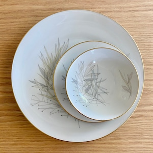 May include: A set of three white ceramic plates with a gold rim and a delicate gray leaf design. The plates are nested together, with the smallest plate on top.
