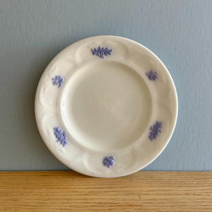 Peut inclure: Une soucoupe en céramique blanche avec un bord festonné et un motif floral dans des tons de violet. La soucoupe est parfaite pour servir du café ou du thé.