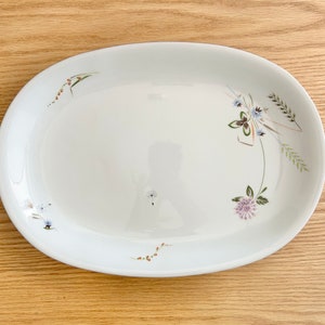 May include: An oval, white serving platter with delicate floral and botanical designs. The platter features a subtle rim and is adorned with clusters of small flowers and sprigs in shades of green, blue, and purple. The design is spread across the surface of the platter.
