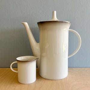 May include: A white ceramic coffee pot and creamer set. The coffee pot has a tall, cylindrical shape with a spout and handle. The creamer is small with a handle. Both pieces have a brown rim and are set against a blue background.