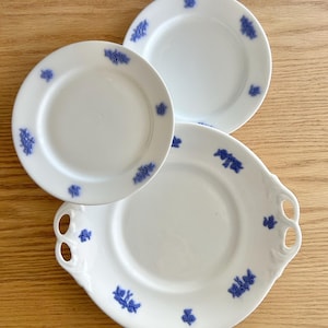 May include: Three white porcelain plates with a blue floral design. The plates are arranged on a light wood surface. One plate has handles. The floral design is repeated around the edges of the plates.