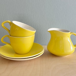 Puede incluir: Un juego de té de cerámica amarillo brillante con dos tazas, dos platillos y una jarra para la crema. Las tazas están apiladas y la jarra tiene un asa curva y un pico. El juego está sobre una superficie de madera clara.
