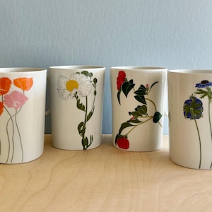 May include: Four white ceramic mugs with floral designs. Each mug features a different flower illustration in shades of orange, pink, white, yellow, red, green, and blue. The mugs have a simple, cylindrical shape with a white handle.