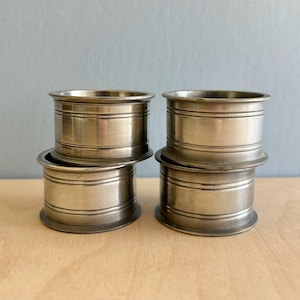 May include: Four silver-colored metal napkin rings stacked in two pairs. Each ring has a cylindrical shape with two horizontal grooves. The rings are arranged on a light-colored wooden surface against a pale blue background.
