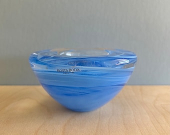 Op de afbeelding: Een blauwe glazen kaarsenhouder met een wit swirl-ontwerp. De houder heeft de tekst "Kosta Boda Since 1942" aan de zijkant.