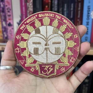 Op de afbeelding: Geborduurde ronde patch in bordeaux, crème en goud. De patch heeft de tekst "THE 'WHEEE' BALANCES THE 'WHOOO'" en het nummer "33". Het centrale ontwerp bevat een gestileerd gezicht.
