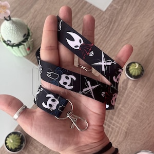 May include: Black lanyard featuring a repeating pattern of white and red character designs. The lanyard has a silver-colored clasp and a matching small strap. The design is set against a black background.