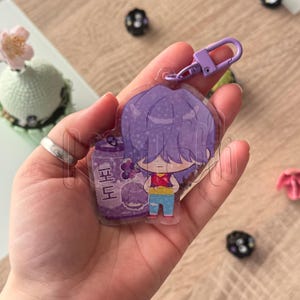 Puede incluir: Un llavero de acrílico transparente con un personaje de dibujos animados de pelo morado, que viste una camiseta roja y pantalones azules, junto a una lata de refresco morada con el texto coreano 포도. Incluye un cierre de mosquetón de color morado.