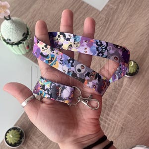 Puede incluir: Un cordón morado con un patrón repetido de fantasmas de dibujos animados y otros personajes espeluznantes. El cordón tiene un cierre de metal plateado y un lazo de tela a juego. El diseño incluye tonos de morado, azul, gris y negro.
