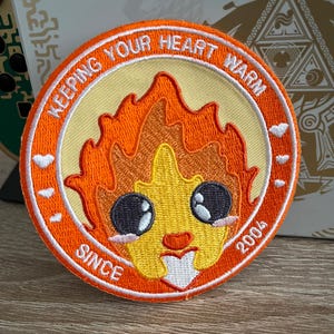 Può includere: Toppa ricamata arancione e gialla con un personaggio di fiamma dei cartoni animati con occhi grandi e un cuore. Il testo intorno al personaggio dice "Keeping Your Heart Warm Since 2004".