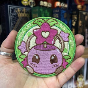 Puede incluir: Parche bordado con un personaje de dibujos animados con cuerpo rosa, ojos grandes y cabeza en forma de flor. El parche tiene un borde verde con las palabras "FAIRYING LIFE BEST LIFE". El diseño incluye detalles en rosa y verde.