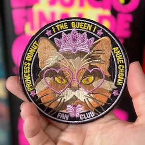 Puede incluir: Un parche bordado circular con un gato calicó que lleva una corona morada y gafas en forma de corazón. El texto dice PRINCESS DONUT, THE QUEEN, ANNE CHONK y FAN CLUB. El diseño incluye pequeñas estrellas rosas y un detalle de mariposa morada.