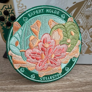 Könnte beinhalten: Ein runder, grüner gestickter Aufnäher mit dem Text "Expert Kolog Collector" am Rand. Die Mitte des Aufnähers zeigt eine rosa Blume mit grünen Blättern und einem hellblauen Hintergrund.
