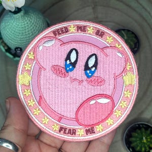 Puede incluir: Parche bordado rosa con un personaje de dibujos animados con ojos grandes y una burbuja rosa. El texto alrededor del personaje dice "Feed Me Or Fear Me".