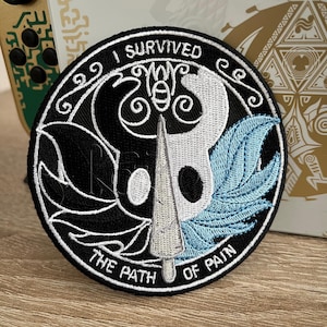 Könnte beinhalten: Schwarzer und weißer gestickter Aufnäher mit einem stilisierten Ritterhelm und einer blauen Feder. Der Text "I Survived The Path Of Pain" ist um den Rand gestickt.