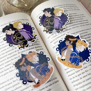 May include: Decorative stickers featuring illustrations of two figures in profile, adorned with crowns and laurel wreaths. The stickers have a dark blue and purple color scheme with gold accents, set against a background of an open book.