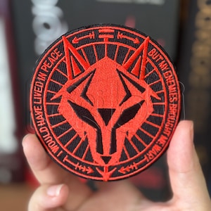 Könnte beinhalten: Ein runder bestickter Aufnäher mit einem stilisierten roten Wolfskopf-Design auf schwarzem Hintergrund. Der Rand enthält den Text I WOULD HAVE LIVED IN PEACE BUT MY ENEMIES BROUGHT ME WAR in roten Großbuchstaben, akzentuiert durch geometrische Linien.