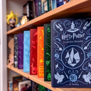 May include: A bookshelf with a row of colorful books, including a blue book with the title "Harry Potter and the Philosopher's Stone" in Spanish. The book has a blue and white illustration of a wizard's hat and a magical symbol.