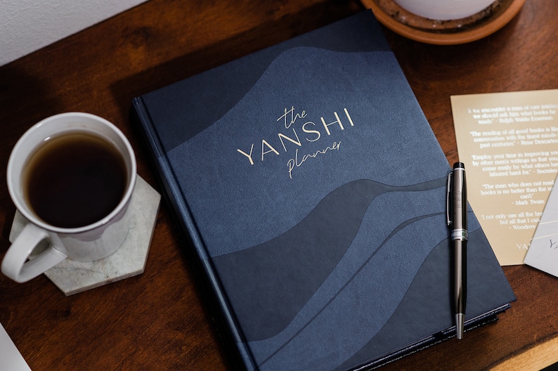 May include: A navy blue planner with gold lettering that reads "the YANSHI planner". The planner is on a wooden surface with a cup of coffee and a pen.
