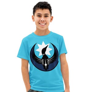 Può includere: Una maglietta azzurra con una grafica di un uccello all'interno di un design a mezzaluna e stella. L'uccello è nero e bianco, e la luna e la stella sono blu e bianche. La maglietta è indossata con pantaloni cargo effetto used e scarpe da ginnastica nere.