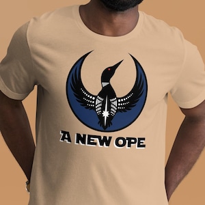 Könnte beinhalten: Beigefarbenes T-Shirt mit einem Design eines schwarz-weißen Vogels in einem blauen Halbmond. Der Text "A NEW OPE" ist darunter gedruckt. Das T-Shirt hat eine neutrale Farbe.
