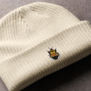 May include: Off-white knitted beanie hat featuring a small embroidered insect design in yellow, orange, and black. The beanie is folded at the brim and rests on a grey surface. The hat has a textured, ribbed knit pattern.