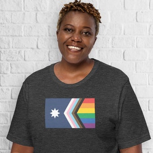 Op de afbeelding: Een donkergrijs t-shirt met een afbeelding van de Progress Pride-vlag, met een witte ster in de linkerbovenhoek.