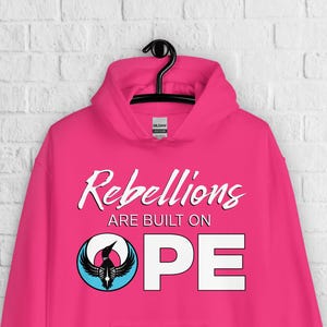 May include: A bright pink hoodie with the words "Rebellions are built on HOPE" in white script and block letters. A circular graphic with a black bird and blue and white accents is centered on the "O" and "P".