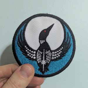 May include: Round embroidered patch featuring a stylized black bird with outstretched wings, a red eye, and a white chest. The bird is set against a white crescent moon within a blue circle, with a black border. The patch is likely for clothing or accessories.