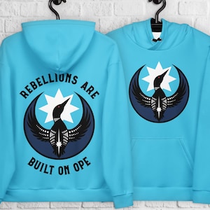 Könnte beinhalten: Zwei hellblaue Kapuzenpullover hängen an schwarzen Haken. Jeder Kapuzenpullover zeigt eine Grafik eines Vogels mit einem Sternmuster in einem dunkelblauen Kreis. Ein Kapuzenpullover hat den Text "REBELLIONS ARE BUILT ON OPE".