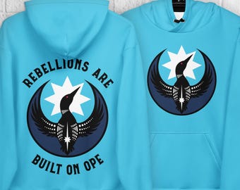 Op de afbeelding: Twee lichtblauwe hoodies hangen aan zwarte haken. Elke hoodie heeft een afbeelding van een vogel met een sterrenpatroon in een donkerblauwe cirkel. Eén hoodie heeft de tekst "REBELLIONS ARE BUILT ON OPE".