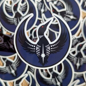 May include: A collection of stickers featuring a stylized black bird with outstretched wings, a white belly, and a red eye, set against a dark blue crescent moon shape. The bird has a star at its chest. The stickers are scattered.