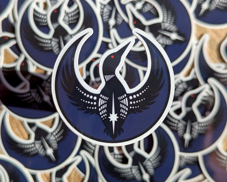 May include: A collection of stickers featuring a stylized black bird with outstretched wings, a white belly, and a red eye, set against a dark blue crescent moon shape. The bird has a star at its chest. The stickers are scattered.