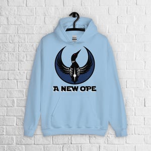May include: Light blue hooded sweatshirt with a black and white bird graphic inside a crescent moon shape. The text "A NEW OPE" is printed below the graphic. The sweatshirt has a front pocket and a drawstring hood.