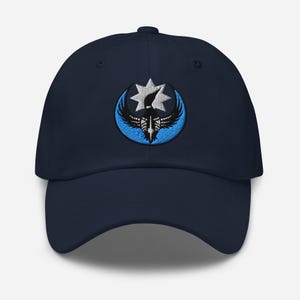 Può includere: Cappellino da baseball blu navy con un emblema ricamato. L'emblema presenta una stella bianca, ali nere e una forma a mezzaluna blu. Il cappellino ha una visiera curva e una chiusura con cinturino in tessuto.