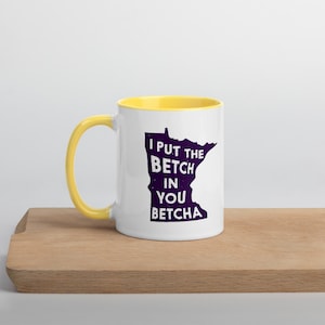 May include: White ceramic mug with a yellow rim and handle. The mug features a purple outline of the state of Minnesota with the text "I put the betch in you betcha".