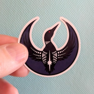 May include: A sticker with a loon design. The loon is black with white details and a red eye, set within a dark blue crescent. The sticker has a white border and is held against a light blue background.