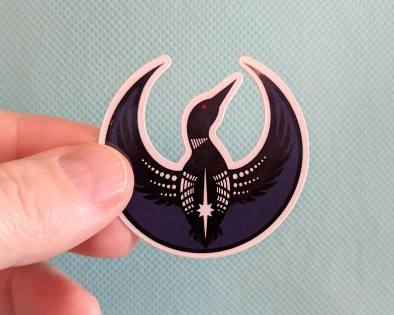 May include: A sticker with a loon design. The loon is black with white details and a red eye, set within a dark blue crescent. The sticker has a white border and is held against a light blue background.
