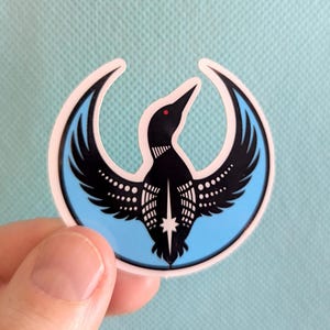 May include: A sticker depicting a black bird with spread wings, set within a light blue crescent shape with a white border. The bird has a red eye and white dot details. The sticker is held against a light blue surface.