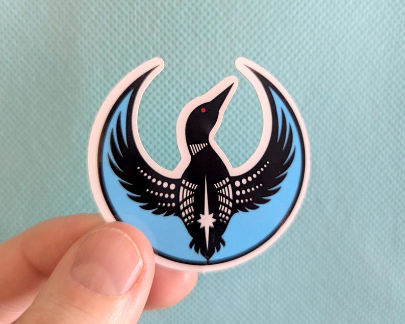 May include: A sticker depicting a black bird with spread wings, set within a light blue crescent shape with a white border. The bird has a red eye and white dot details. The sticker is held against a light blue surface.